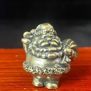 May include: A small, solid brass figurine of Santa Claus with a textured hat, a thick beard, and a sack held in one hand. The figure features a polished metallic finish with darkened recessed areas that highlight the sculpted details of the clothing.