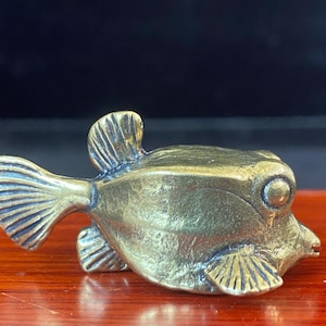 May include: A small, metallic brass figurine shaped like a boxfish. The object features a textured, golden-brown finish with detailed, etched lines on the fins and a prominent, circular eye. The body is compact and angular, capturing the distinct form.