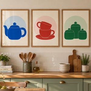 May include: Three framed wall art prints featuring kitchenware illustrations. The left print shows a blue teapot on a light blue circle. The centre print displays two red stacked cups on a pink circle. The right print depicts three green jars on a light green circle.