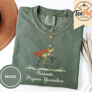 May include: A moss green crew neck t-shirt featuring a graphic of a frog wearing a gold crown and a red cape while riding a penny-farthing bicycle. Below the frog, the text reads, Peasants, Prepare Yourselves. The shirt has a Comfort Colors label.