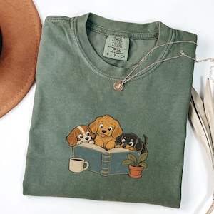 May include: An olive green crew neck t-shirt featuring a graphic of three cartoon dogs reading an open book. The print includes a small potted plant and a coffee mug. The shirt has a Comfort Colors brand label at the neck indicating size small.