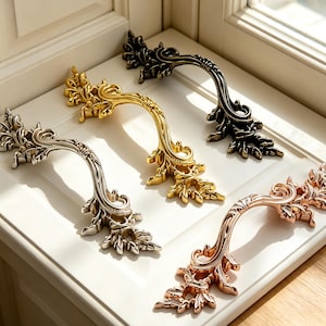 May include: Four ornate metal drawer pulls arranged in a diagonal line. Each handle features intricate leaf and scrollwork detailing. The finishes include a polished silver, a bright gold, a dark antique bronze, and a warm rose gold metallic tone.