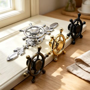 May include: A collection of four cabinet knobs shaped like ship steering wheels, featuring a polished silver finish with an ornate backplate, a brushed bronze finish, a shiny gold finish, and a matte black finish, all mounted on a white panel.