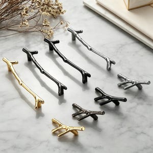 Può includere: Una collezione di maniglie per mobili a forma di rami d'albero, che comprende tiranti lunghi e pomelli più piccoli. La ferramenta è disponibile in finiture oro metallizzato, nero opaco e argento spazzolato, con un aspetto organico.