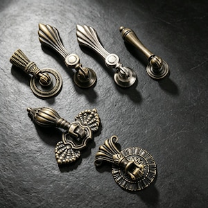 May include: A collection of six antique brass-toned metal furniture pulls and drawer handles. The set includes various shapes such as ring pulls, drop handles, and ornate backplates with intricate ribbed, scalloped, and rope-like textured details.