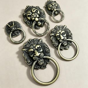 Pode incluir: Um conjunto de cinco puxadores de gaveta em metal com tom de latão antigo, apresentando designs de cabeça de leão com argolas circulares, além de um puxador menor com motivo de cabeça de dragão, todos com detalhes em relevo e acabamento metálico envelhecido.