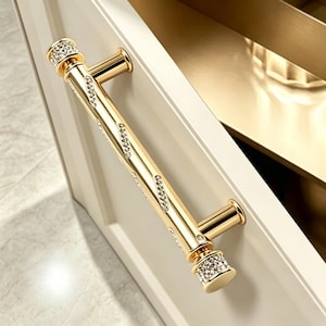 May include: A gold-toned cabinet handle featuring a cylindrical bar with diagonal rows of embedded clear crystals and matching crystal-encrusted circular end caps, mounted horizontally on a cream-colored cabinet door surface.