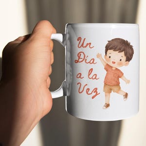 Puede incluir: Una taza de cerámica blanca con una ilustración estilo acuarela de un niño pequeño de cabello castaño que viste una camiseta color óxido y pantalones cortos beige. El texto "Un Día a la Vez" aparece en letra cursiva marrón rojiza.