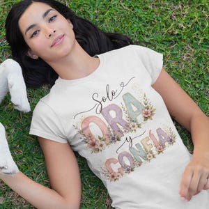 Puede incluir: Una camiseta de manga corta color crema con el texto Solo ORA y CONFIA en letras grandes de tonos apagados decoradas con ilustraciones florales. El diseño incluye un pequeño corazón sobre la palabra Solo y detalles botánicos alrededor.