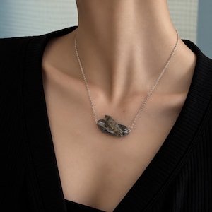 Op de afbeelding: Een zilverkleurige ketting met een hanger bestaande uit twee ruwe, donkergrijze en doorschijnende kristalpunten die met elkaar zijn verbonden. De ketting rust op het sleutelbeen en toont een metalen ketting en een natuurlijke steentextuur.