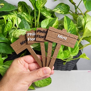 Peut inclure: Quatre marqueurs de plantes marron avec une finition texturée imitation bois. Chaque marqueur présente des lettres blanches en relief. Les étiquettes indiquent Mint, Philo et Flor. Ces panneaux rectangulaires sont fixés sur des tiges fines.