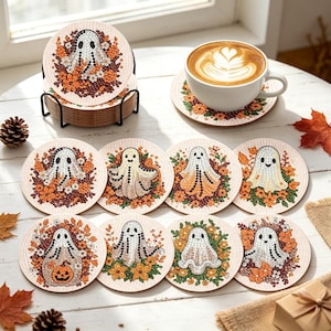 May include: A set of round drink coasters featuring diamond art designs of ghosts surrounded by autumn flowers and leaves. The coasters are displayed in a black metal holder, with one coaster placed under a white ceramic mug of latte.