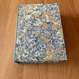 May include: A hardback book featuring a marbled paper cover with a swirling pattern of mustard yellow, forest green, white, and dark blue colours. The spine of the book is covered in a solid light blue material, providing a clean contrast.