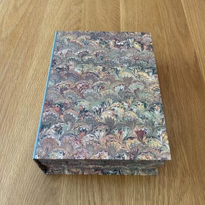 May include: A rectangular book with a hard cover featuring a marbled pattern in shades of sage green, muted red, cream, and dark blue. The spine of the book is a solid light blue colour, and the cover displays a repeating fan-like design.