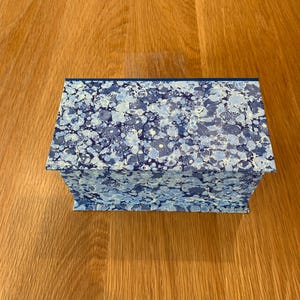 May include: A rectangular box featuring a marbled pattern in various shades of blue, light blue, and white. The box has a flat lid and a structured design, with the intricate, swirling marbled paper covering all visible exterior surfaces.