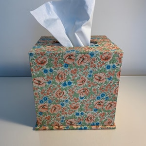 May include: A square tissue box cover featuring a dense floral pattern with peach-coloured blossoms, small blue flowers, and green foliage. A single white tissue is pulled through the circular opening at the top of the box.
