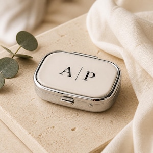 May include: A rectangular metal pill case with a polished silver finish and a smooth cream-colored lid. The top of the lid features the black printed letters A | P centered in a serif font. The case has a hinged lid and a small latch mechanism.