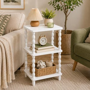 May include: A three-tier white side table featuring four spindle-style legs with a rounded, stacked bead design. The table has three square shelves, with the top shelf holding a small lamp, a potted plant, and a candle, and the middle shelf holding books.