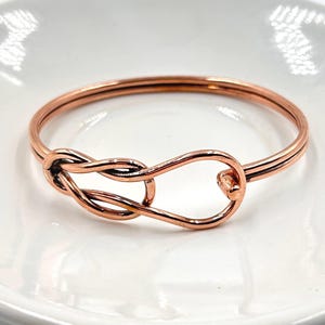 Peut inclure: Un bracelet jonc en métal de couleur cuivre poli présentant un design en fil continu formant un nœud de style infini au centre. Le bracelet possède une surface lisse et réfléchissante ainsi qu'une forme circulaire avec une fermeture à crochet.
