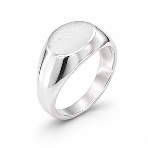 Könnte beinhalten: Ein polierter Siegelring aus Silber mit einer glatten, abgerundeten Ringschiene und einer flachen, ovalen Oberseite mit texturierter, matter Oberfläche. Das Metall reflektiert das Licht und betont den Kontrast zwischen dem glänzenden Ringband.
