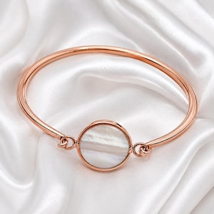 May include: A rose gold bangle bracelet featuring a central circular charm inlaid with a smooth, iridescent white mother-of-pearl disc. The metal band is thin and polished, connecting to the circular centerpiece with small, delicate metal loops.