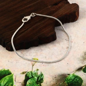 May include: A thin, silver-coloured snake chain bracelet featuring a polished metallic finish and a secure lobster claw clasp. The bracelet is formed into a circular shape, showcasing the smooth, interlocking links of the chain design.