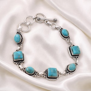 May include: A silver-toned metal bracelet featuring alternating oval and square-shaped turquoise stones set in decorative bezels. The bracelet is secured with a toggle clasp consisting of a circular ring and a T-bar closure, connected by silver links.