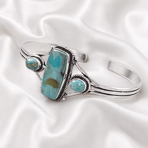 May include: A silver-toned cuff bracelet featuring a large rectangular turquoise stone in the centre flanked by two smaller oval turquoise stones. The metal band has a twisted rope texture around each stone and a triple-line design on the sides.