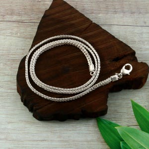 Puede incluir: Un collar de cadena tipo trigo en tono plateado con un diseño de eslabones metálicos trenzados y ajustados. El collar está terminado con un cierre de langosta pulido en tono plateado en un extremo y un pequeño conector cilíndrico en el otro.