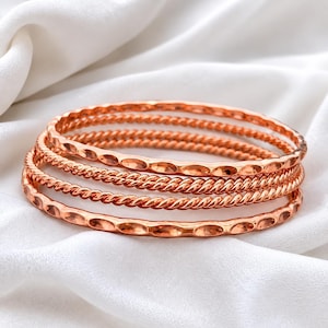 May include: A set of four stacked bangles in a metallic rose gold tone. The set includes two bangles with a smooth, hammered texture and two bangles featuring a twisted, rope-like design. Each bangle is circular and maintains a consistent finish.