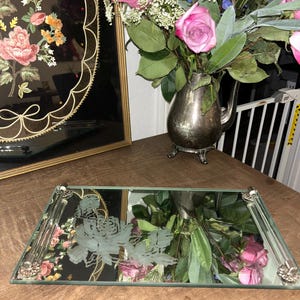 Esta bandeja de tocador de estilo vintage cuenta con una superficie de espejo rectangular con detalles florales grabados y asas decorativas de metal.