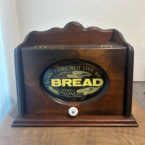 May include: A dark brown wooden bread box with a hinged front door featuring an oval glass window. The glass displays the yellow text "MAN DOES NOT LIVE BY BREAD ALONE" with wheat illustrations. A small white ceramic knob is centered on the door.