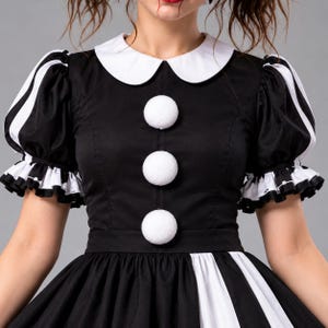 Puede incluir: Un vestido en blanco y negro con cuello redondo blanco, tres grandes botones de pomp&oacute;n blancos en el centro y mangas cortas abullonadas con pu&ntilde;os de volantes blancos y negros. El corpi&ntilde;o es negro liso y la falda tiene rayas verticales.