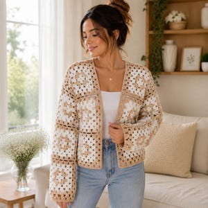 Pode incluir: Um cardigã de crochê de manga comprida com um design de quadrados em fio bege e branco. A peça aberta na frente é usada sobre uma blusa branca lisa e calças jeans azul-claras, exibindo uma construção de malha artesanal texturizada.
