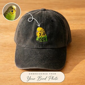 Op de afbeelding: Een antracietgrijze baseballpet met een gedetailleerde geborduurde patch van een gele en groene grasparkiet. Een witte handgetekende pijl wijst vanuit een ronde inzetfoto van dezelfde vogel naar het geborduurde ontwerp op het voorpaneel.