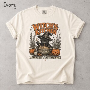 May include: An ivory short-sleeve t-shirt featuring a graphic of a black cat wearing a witch hat, stirring a bowl labeled Magic Cookie Dough. The text reads Witch's Kitchen and Made With A Little Magic, surrounded by pumpkins and herbs.
