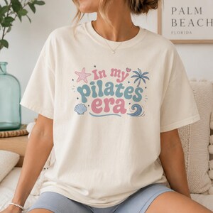 May include: A cream-colored short-sleeve t-shirt featuring the text "in my pilates era" in blue and pink retro-style lettering. The design includes graphic illustrations of a pink starfish, a blue palm tree, a blue seashell, and a blue wave.