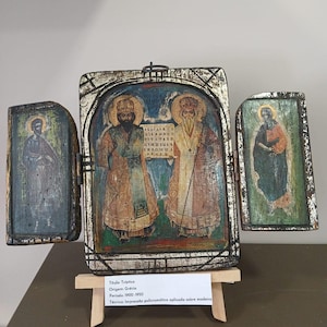 Puede incluir: Un tríptico de madera con figuras religiosas vestidas con túnicas y aureolas. El panel central muestra a dos figuras sosteniendo un pergamino con texto, mientras que los paneles laterales representan figuras individuales. Marco con acabado plateado envejecido.