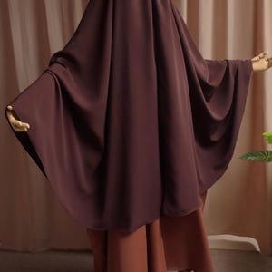 May include: A dark brown, floor-length, loose-fitting garment with a matching head covering that drapes over the shoulders and arms, worn over a lighter, terracotta-colored skirt. The ensemble features a solid, matte fabric with wide sleeves.