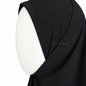 Può includere: Un hijab nero a tinta unita drappeggiato su una testa di manichino bianca. Il tessuto presenta una finitura liscia e opaca con cuciture visibili lungo il bordo anteriore. Il testo Modesty Tailor Studio è stampato in piccole lettere grigio chiaro sul lato.