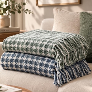 May include: Two folded throw blankets stacked on a sofa. The top blanket features a green and white checkered pattern with fringe edges. The bottom blanket features a blue and white checkered pattern with fringe edges. Both blankets have a textured weave.