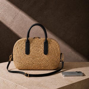 May include: A structured handbag crafted from woven natural straw with a rounded rectangular shape. It features black leather handles and a matching adjustable black shoulder strap with gold-toned metal hardware, including a top zipper closure.
