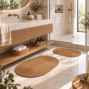 May include: Two oval-shaped, woven jute bath mats in a natural tan color are placed on a light-colored tiled floor. The mats feature a concentric circular pattern with a tight, braided texture and a small dark brown leather-like tag on the edge.