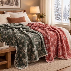 May include: Two plush throw blankets draped over a bed, one in dark green and one in red, both featuring a repeating white snowflake pattern. The blankets have a soft, textured appearance with visible white trim along the edges of the fabric.