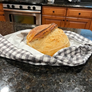 May include: A round loaf of crusty sourdough bread with a golden-brown exterior and a distinct scored top, resting in the center of a white and grey checkered cloth napkin. The bread has a rustic texture and a visible rise from the baking process.