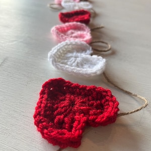 Puede incluir: Una guirnalda de corazones de ganchillo hechos a mano en tonos rojos, rosas y blancos. Los corazones están ensartados en un hilo natural, creando una pancarta decorativa. Ideal para el Día de San Valentín u otras ocasiones especiales.