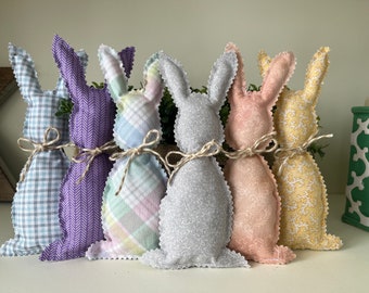 May include: Six fabric bunny decorations in pastel colors. Each bunny has a unique patterned fabric, including plaid, herringbone, and floral designs. They are tied with twine bows and have a scalloped edge.