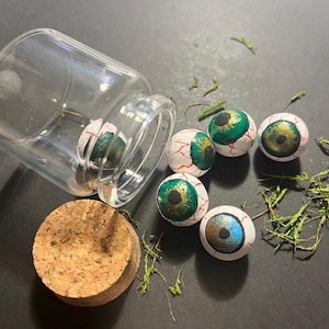 Könnte beinhalten: Ein klares Glasgefäß mit einem Korken, umgeben von mehreren bemalten Augapfeldekorationen. Die Augäpfel sind weiß mit roten Adern, grünen und goldenen Iris und einer blauen Iris auf einem. Grünes Moos sorgt für einen natürlichen Touch.
