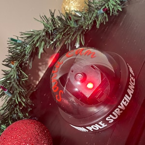 以下が含まれることがあります： 赤いライトが点灯した黒と白のサンタカムポール監視カメラ。赤いオーナメントと金のオーナメントなど、クリスマスの飾りで囲まれている