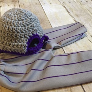 May include: Gray and purple crocheted baseball cap with a matching pair of gray and purple striped pants.